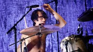 Schlagzeuger mit nacktem Oberkörper vor lila Hintergrund.