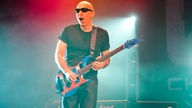 Joe Satriani spielt auf seiner Gitarre und reißt dabei den Mund auf