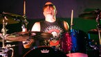Der Drummer von Chickenfoot sitzt an seinem Schlagzeug und schwingt seine Sticks. Erträgt eine braune Sonnenbrille und hat eine Glatze