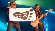 Sammy Hagar hält einen Banner mit der Aufschrift `That shit rocks`rechts daneben Michael Anthony an seinem Bass