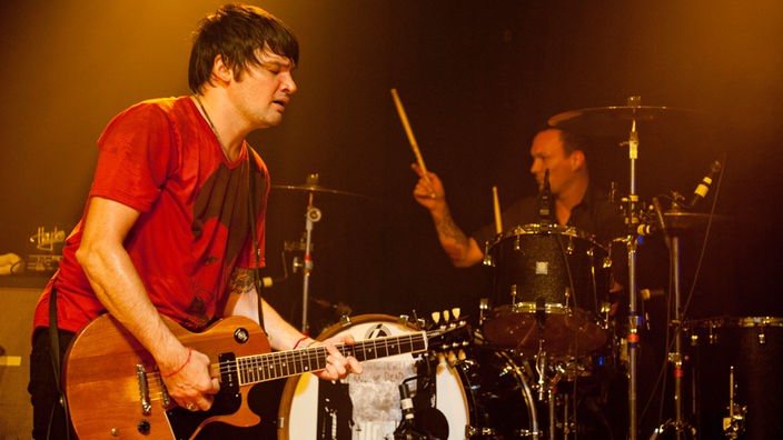 Mann im roten Shirt spielt mit geschlossenen Augen Gitarre, im Hintergrund spielt der Drummer Schlagzeug