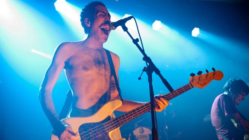Bassist mit nacktem Oberkörper spielt Bass und singt mit weit offenem Mund in ein Mikrofon im blauen Licht.