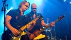 Bassist und Gitarrist der Band "The Chuck Norris Experiment" spielen nebeneinander im blauen Bühnenlicht.