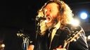 Ein Mann mit langen Haaren und Bart guckt böse und singt in ein Mikrofon.