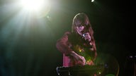 Frau im roten Kleid dreht an einem Syntesizer und singt dabei in ein Mikrofon im hellen Bühnenlicht
