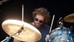 Drummer mit Pilotenbrille, brauen Locken und Vollbart spielt Schlagzeug