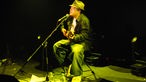 Der Musiker Martin Jondo sitzend und in grünes Scheinwerferlicht getaucht während seines Auftritts auf der Bühne im Underground Köln
