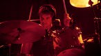 Schlagzeuger trägt beim Spielen Kopfhörer. Die Bühne ist rot beleuchtet.