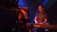 Im Vordergrund ist eine große Kamera bei Bootleg im April 2006 zu sehen, hinter welcher der Frontmann von Rivo Drei an seinem Keyboard steht und singt.