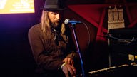 Der Keyboarder der Formation sitz vor seinem Instrument und sieht dabei auf seine Finger. Er hat lange glatte Haare, einen Bart und trägt ein braunes Hemd und einen schwarzen Hut.