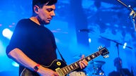Der Gitarrist von Rakede steht vor einer blau erleuchteten Bühne und sieht angestrengt auf seine Gitarre.