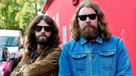 Zwei Bandmitglieder der Band "The Sheepdogs" stehen vor einer roten Wand. Beide haben einen langen Bart, lange Haare und tragen dunkle Sonnenbrillen.
