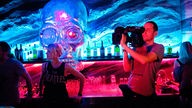 Ein Kameramann steht mit voller Ausrüstung an der Bar und ist am Filmen. Im Hintergrund sieht man einen riesigen gläsernen Schädel an der Wan hängen dessen eines Auge rot erleuchtet ist.