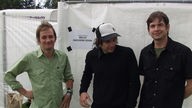Drei Bandmitglieder der Band Wilco stehen vor einem Bauzaun.