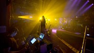 Ein Foto aus der Sicht der Band, von der Bühne in Richtung Publikum, ein gelber Lichtstrahl blendet und malt die Umrisse der Banjospielerin der Band Honig