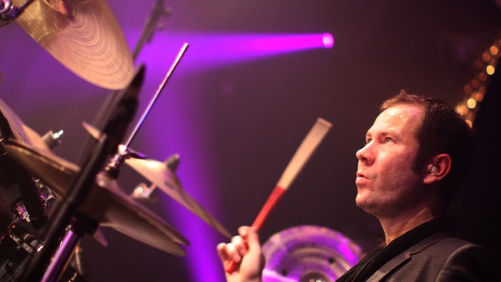 Damon Sawyer am Schlagzeug: Er hält einen Stick mit der rechten Hand hoch und schaut nach oben