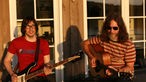 Pink Mountaintops beim Interview sitzen vor einer Terassentür im Sonnenuntergang, Bassist mit Rickenbackerbass zur linken und Gitarrist und Sänger mit Akustikgitarre zur rechten