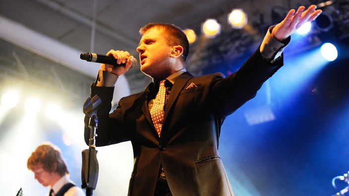 Junger Mann mit Anzug gestikuliert und singt in ein Mikrofon.