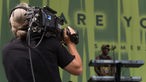Ein WDR-Kameramann filmt, im Hintergrund verschwommen zu erkennen: Demar Gayle, Keyboarder von Raging Fyah, and seinem Instrument.
