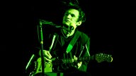 Gitarrist steht vor einem Standmikrofon und wird von grünem Licht bestrahlt