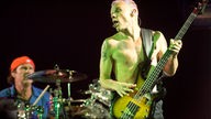 Bassist mit nacktem Oberkörper und Bass spielend streckt die Zunge heraus und guckt nach rechts; im Hintergrund ein Schlagzeuger und das Schlagzeug
