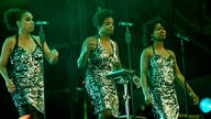 Drei dunkelhäutige Frauen mit schwarzem Haar in jeweils einem hell glitzerndem Kleid singen in jeweils ein Mikrofon