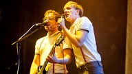 Gitarrist und Sänger stehen beieinander und singen in jeweils ein Mikrofon; dabei werden sie von gelbem Licht bestrahlt