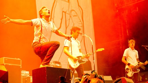 Drei Männer mit weißem Shirt und dunkler Hose auf der Bühne; einer steht mit einem Bein auf einen Verstärker und breitet die Hände aus, einer spielt E-Gitarre, einer spielt E-Bass
