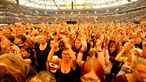 Stadion von innen, es ist gefüllt mit jubelenden Menschen, die ihre Hände in die Luft erheben