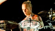 Schlagzeuger mit langen zu einem Zopfg gebundenen Haaren, nacktem Oberkörper und vielen Tatoos auf dem Körper spielt mit einem ernsten Gesichtsausdruck ein Schlagzeug