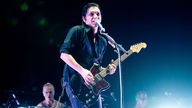 Gitarrist in schwarzem Hemd und mit schwarzen Haaren spielt eine rötlich schimmernde Gitarre und singt in ein Mikrofon, im Hintergrund ist ein Schlagzeuger zu erkennen
