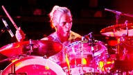 Tätowierter Mann spielt mit ernstem Gesichtsausdruck Schlagzeug, das Bild ist in Rot- / Pinktönen gehalten