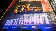 Man sieht eine Leinwand, auf der "1Live Rock im Pott" projeziert wird, darüber ein Bildschirm mit einem Mann am Schlagzeug; im Hintergrund sieht man das Gerüst der Bühne