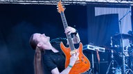 Der Gitarrist der Band lehnt sich nach hinten und streckt seine orangene Stratocaster-Gitarre in die Höhe