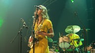 The Zutons: Frau im gelben Kleid steht mit einem Saxophon auf der Bühne