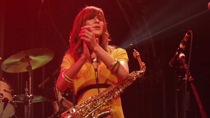 The Zutons: Eine Frau im gelben Kleid hat ein Saxophon umgehängt und klatscht im roten Bühnenlicht