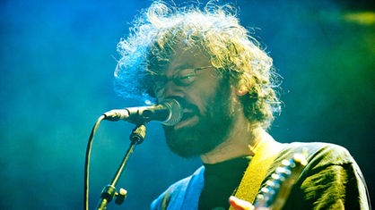 Der Sänger und Gitarrist von Notwist steht in gelb-blauem Licht an seinem Mikrofon.