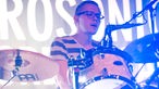 Schlagzeuger mit Brille und gestreiftem Shirt singt in ein Mikrofon im blau-weißen Bühnenlicht