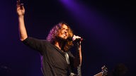 Der Sänger von Soundgarden hält sich mit der linken Hand am Mikrofon fest und streckt die Rechte in die Höhe.