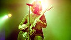 Der Frontmann steht singend und Gitarre spielend in gleißend grünem Licht vor dem Mikrofon.