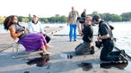 Sängerin Joy Denalane und das Kamera-Team bereiten sich auf ein Interviw am Fühlinger See vor. Denalane sitzt in einem Stuhl, vor ihr kniet das Team und prüft die Kameraeinstellung
