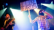 Der Sänger hält ein Schild hoch auf welchem steht " I Like Beards"