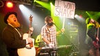 Gitarrist, Bassist und der Keyboarder mit einem Schild, auf dem "I Wont Shave" steht, in der Hand.