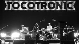 Bandfoto: Tocotronic bei einem Live-Auftritt 2006; schwarz-weiss Bild der vier Musiker auf der Bühne; im Zentrum das Schlagzeug umsäumt von Lautsprechern und Mikrofonständern