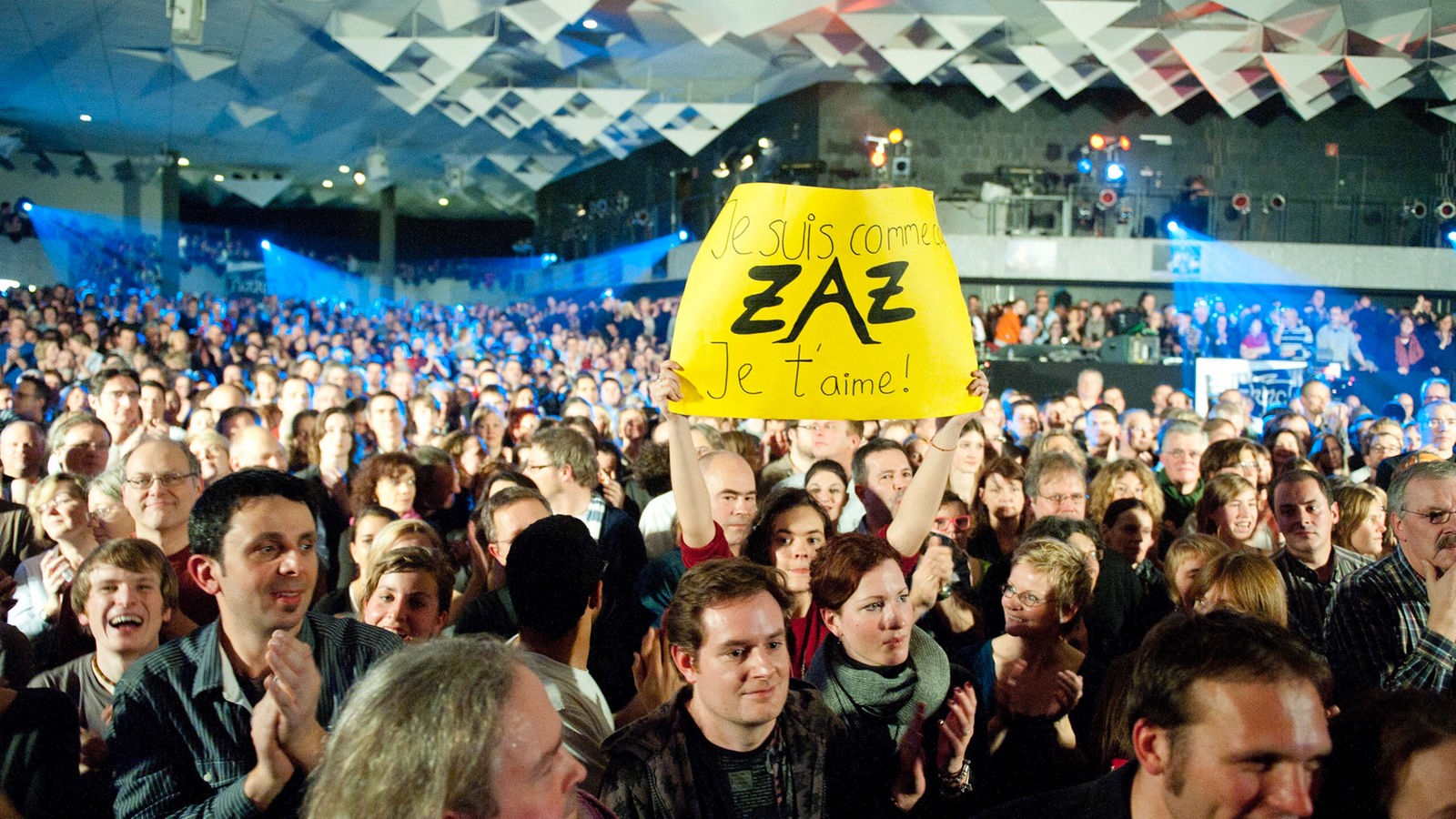 Mit Menschen gefüllte Halle, ein Mädchen hält ein gelbes Schild hochhält, auf dem steht "Je suis comme ZAZ Je t'aime!"
