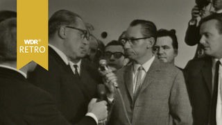 Herbert Wehner steht umringt von einer Gruppe neben einem Reporter und spricht in ein Mikrofon.