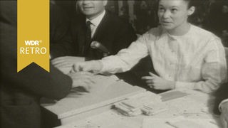 Ein Mann, der steht, gibt einen Stimmzettel ab. Am Tisch ihm gegenüber sitzt eine Frau, die ihn anblickt.