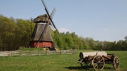 Auf dem Bild steht eine Windmühle. Im Vordergrund steht ein Karren, der einen Holzstamm transportiert.