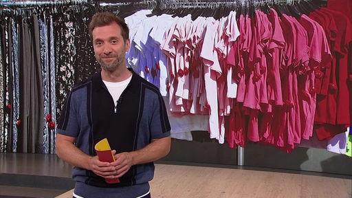  Daniel Aßmann moderiert im Servicezeit-Studio. Im Hintergrund sind Kleiderstangen anbgebildet.