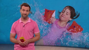 Daniel Aßmann steht im Servicezeit-Fernsehstudio. Hinter ihm sieht man ein kleines Mädchen im Schwimmbad.